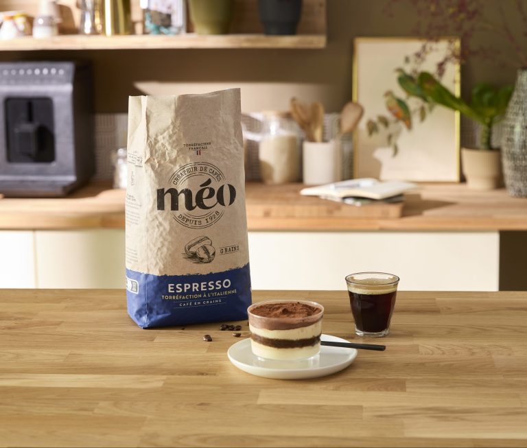 Un sachet de grains de café Méo Espresso, un verre de café expresso onctueux et une assiette avec un dessert bio sont posés sur un plan de travail en bois dans une cuisine.